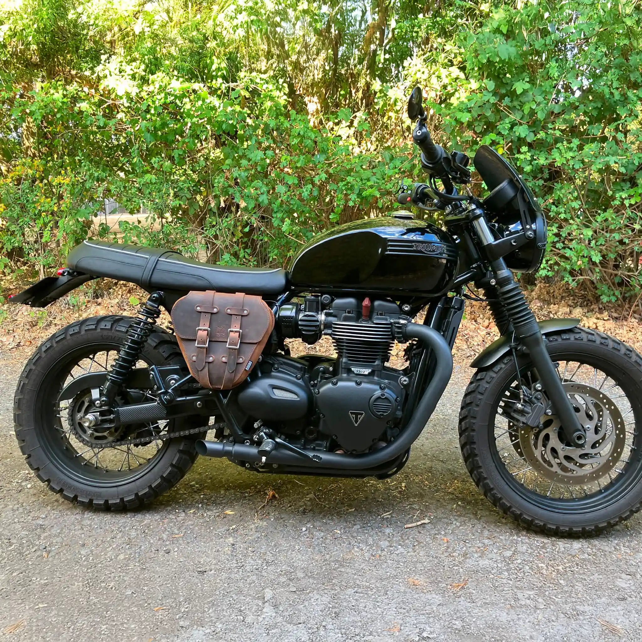 A black Triumph motorcycle with a brown leather saddlebag pouch, parked outdoors on a gravel path with green foliage in the background.